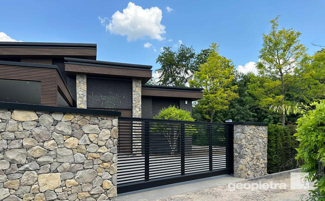 Entrance of a luxurious modern villa with perimeter walls and facades clad in Geopietra manufactured stone, modern gate, and surrounding greenery.
